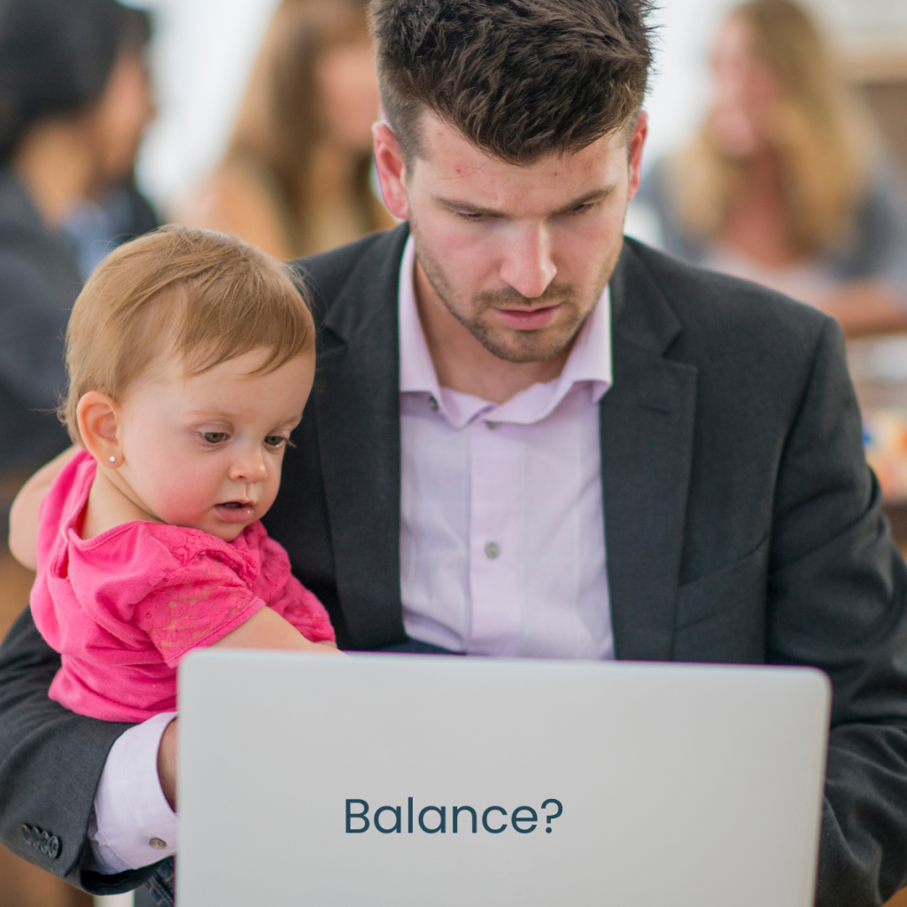 Dad working at laptop while holding a baby in one arm.